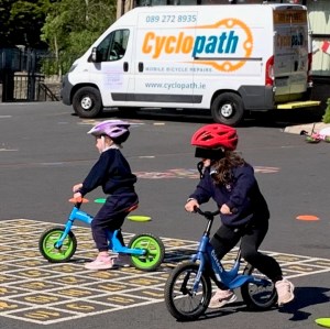 Two children riding balance bikes on a playground surface, wearing protective helmets, with a visible Cyclopath bicycle repair van in the background.